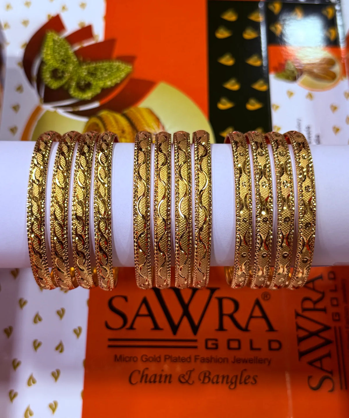 A set of gold-plated bangles with intricate floral and leaf patterns, displayed on a white cylindrical stand. The background features branded packaging for SAWRA GOLD.