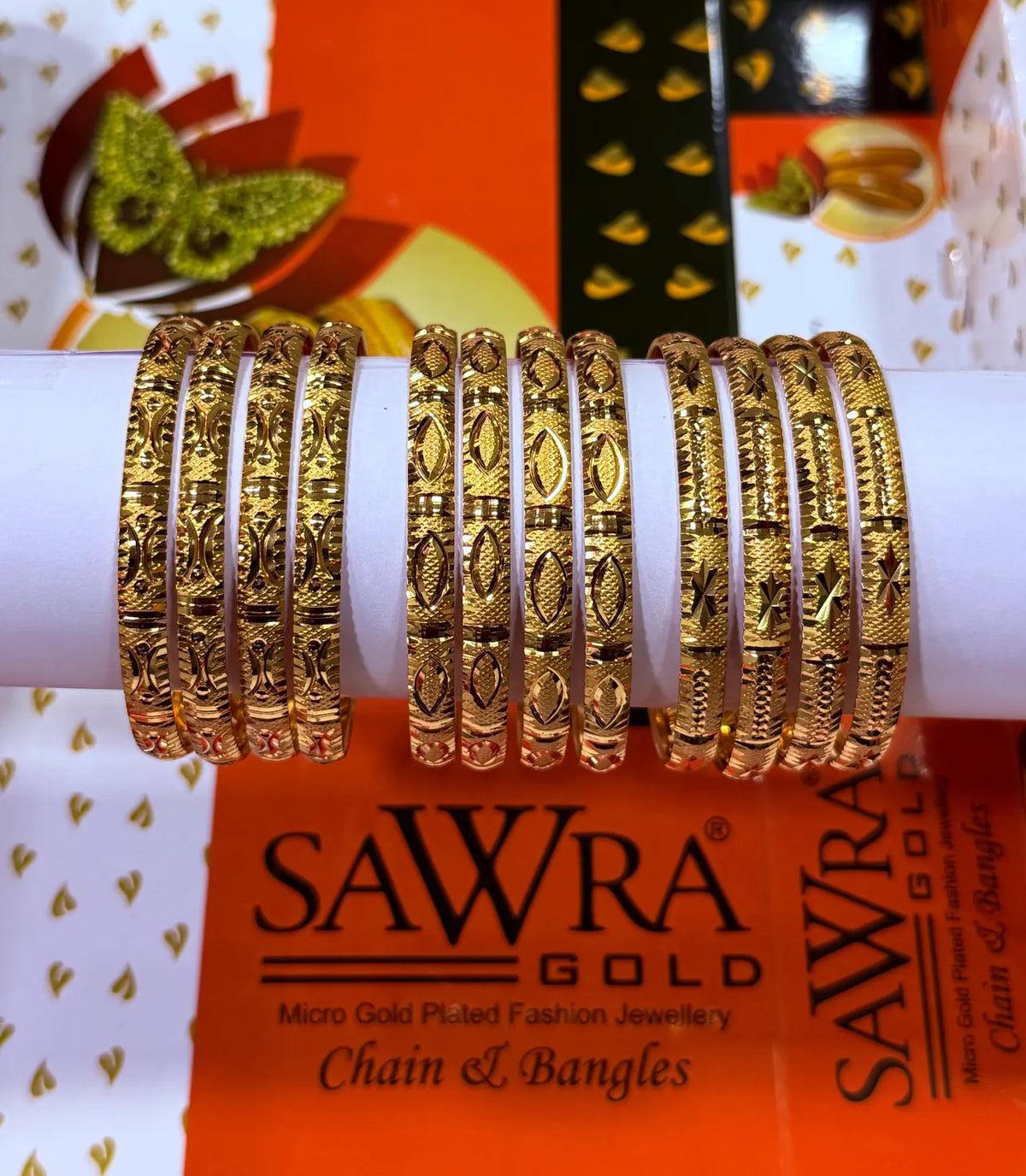 A set of gold-plated fashion bangles with intricate floral and geometric engravings, displayed on a white cylindrical stand. The background features branded orange and white packaging for SAWRA GOLD.