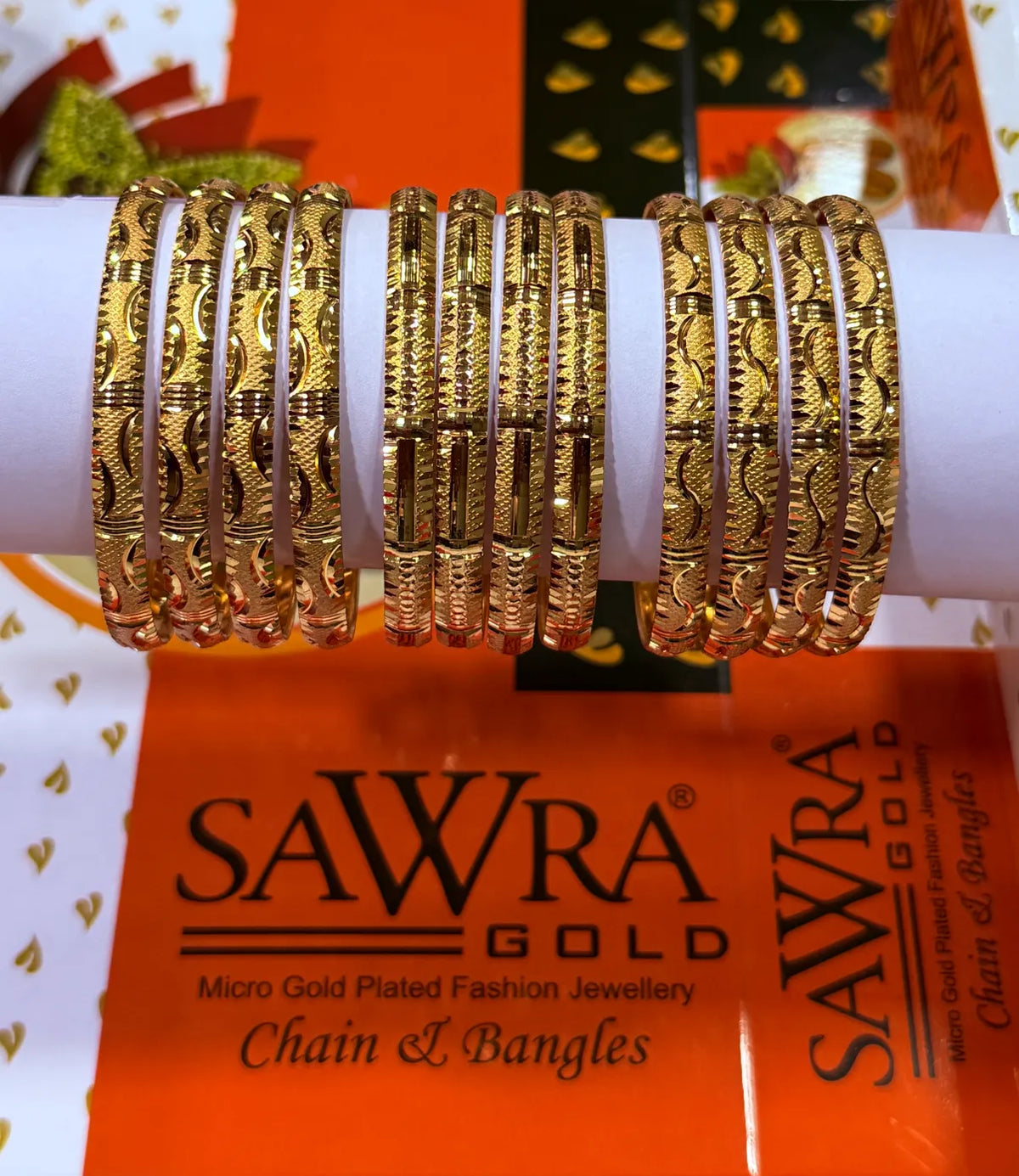 A set of gold chain and bangle fashion jewellery displayed on a white cylindrical stand, with an orange SAWRA GOLD branded box underneath.