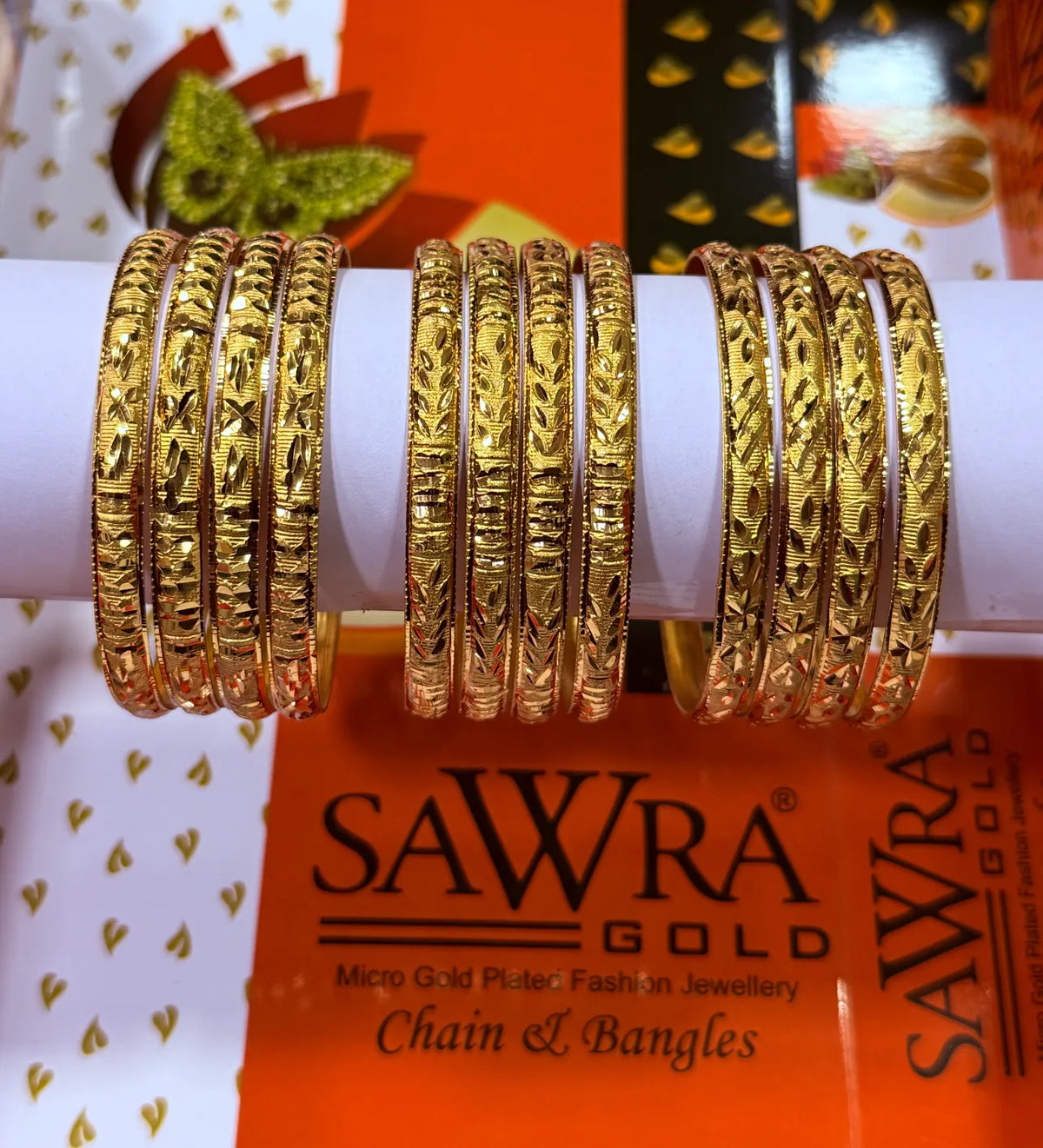 Three sets of gold-plated bangles with intricate floral and geometric patterns, displayed on a white cylindrical stand. The background features the orange and white packaging of Sawra Gold chain and bangle set.