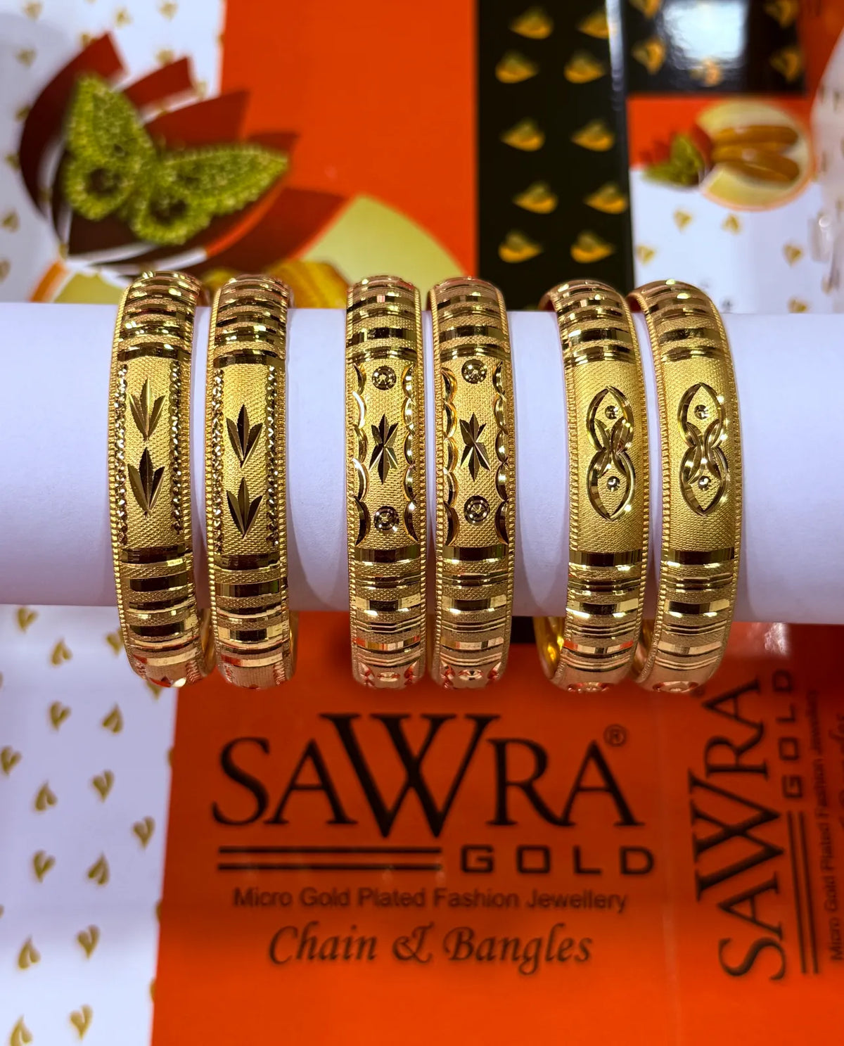 Five gold-plated bangles with intricate floral and geometric engravings, displayed on a white cylindrical stand. The background features the brand packaging for SAWRA GOLD.