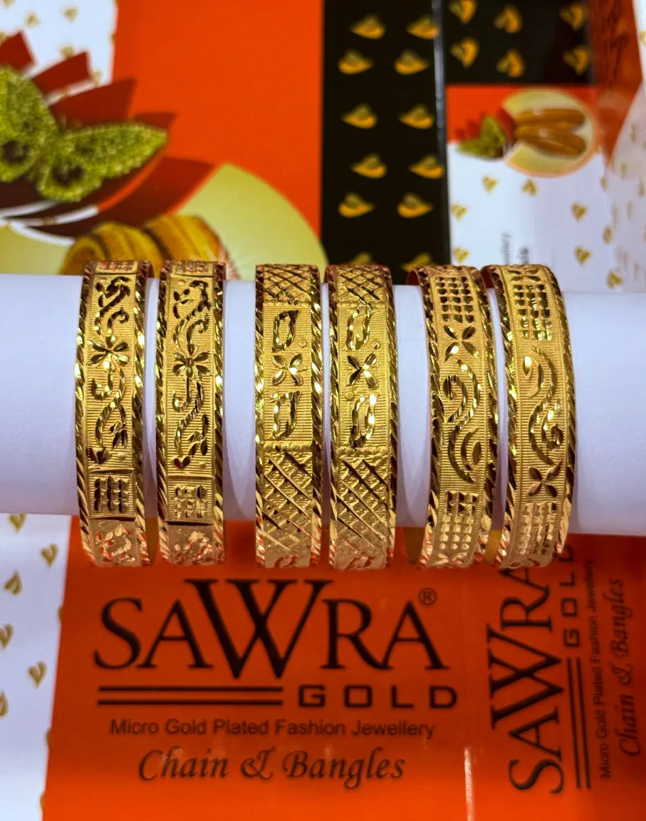 Five ornate gold-plated chain and filigree bangles displayed on a white cylindrical stand, with an orange SAWRA GOLD brand box visible underneath.