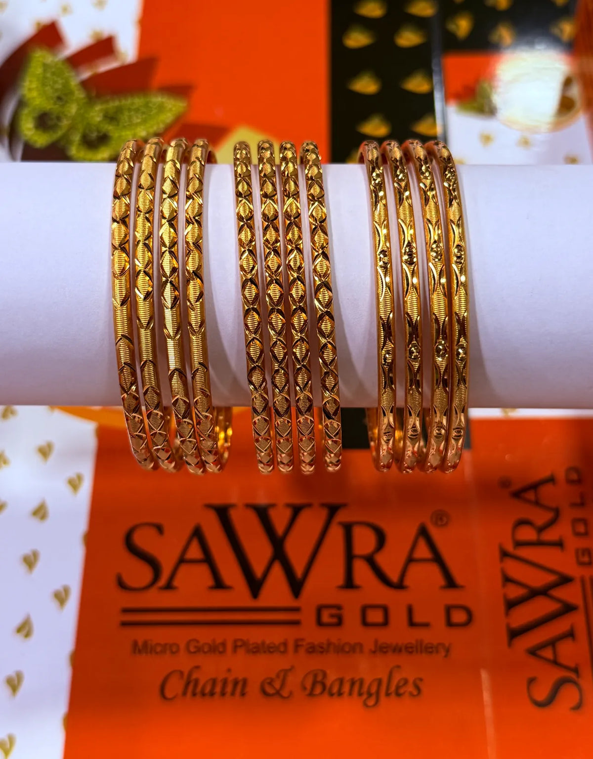 A set of gold-plated bangles displayed on a white cylindrical stand, with SAWRA GOLD branding visible on the packaging below.