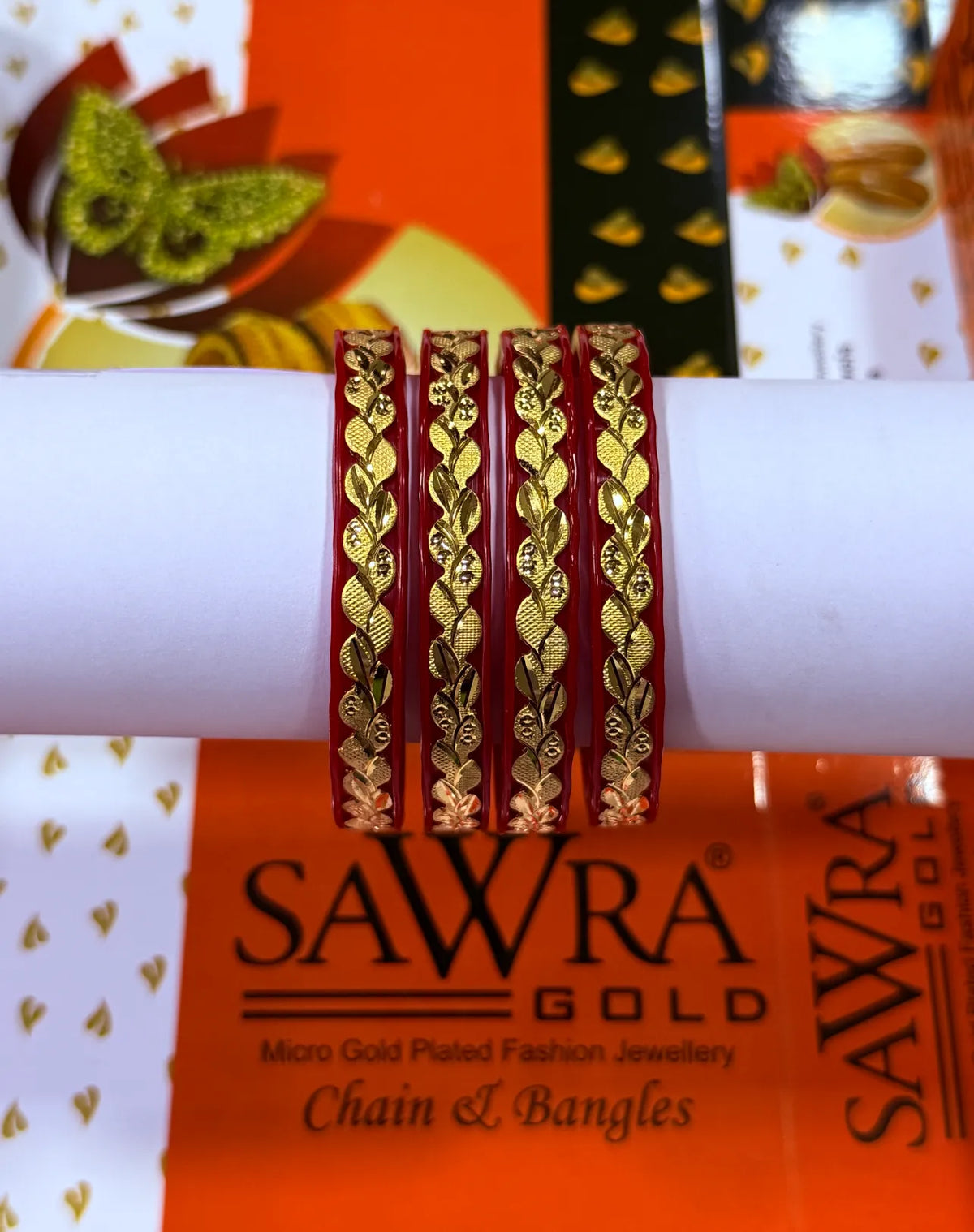 Four red bangles with gold leaf-patterned borders wrapped around a white cylindrical display, set against a branded orange and white Sawra Gold background.