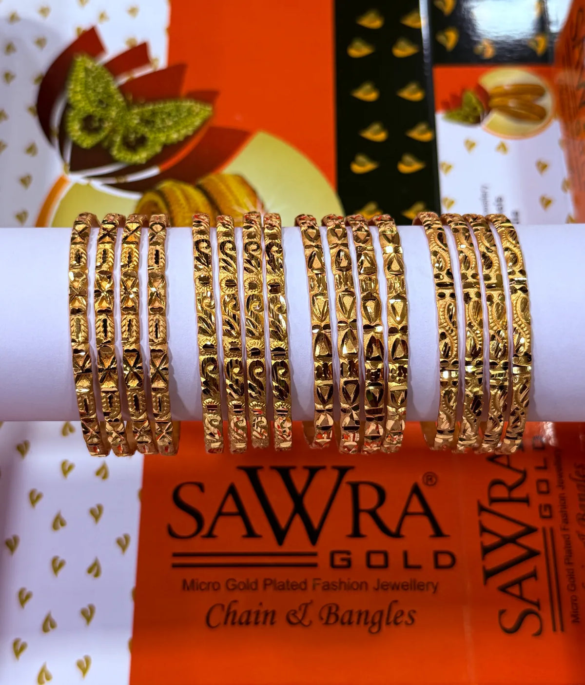 A set of gold chain and bangle necklaces displayed on a white cylindrical stand, with SAWRA GOLD branding visible on the packaging in the background.