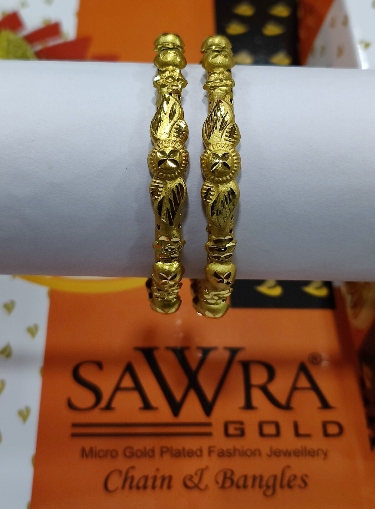 Gold bangles on a white cylindrical object with 'Sawra Gold' branding in the background.
