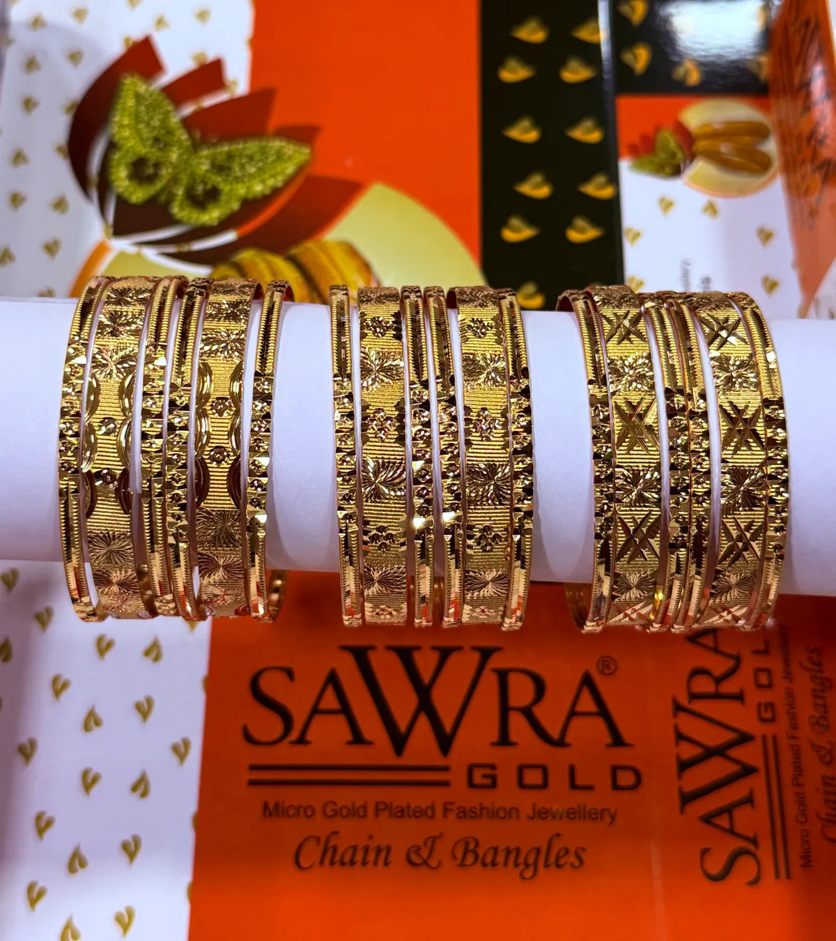 Four stacks of gold-plated bangles with intricate floral and geometric patterns displayed on a white cylindrical stand, set against a branded orange and white SAWRA GOLD background.