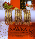 A set of gold-plated fashion bangles displayed on a white cylindrical stand, with SAWRA GOLD branding visible on orange packaging in the background.