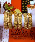 Six ornate gold bangles with intricate engraved patterns displayed on a white cylindrical stand, with a SAWRA GOLD brand box visible in the background.
