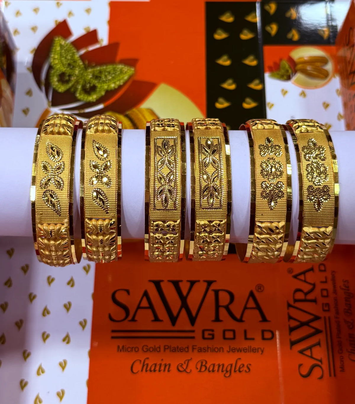 Six gold chain link bangles displayed on a white cylindrical stand, with SAWRA GOLD branding visible in the background.