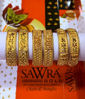 Six ornate gold-plated bangles displayed on a white cylindrical stand, with a branded orange and white SAWRA GOLD chain and bangles box in the background.
