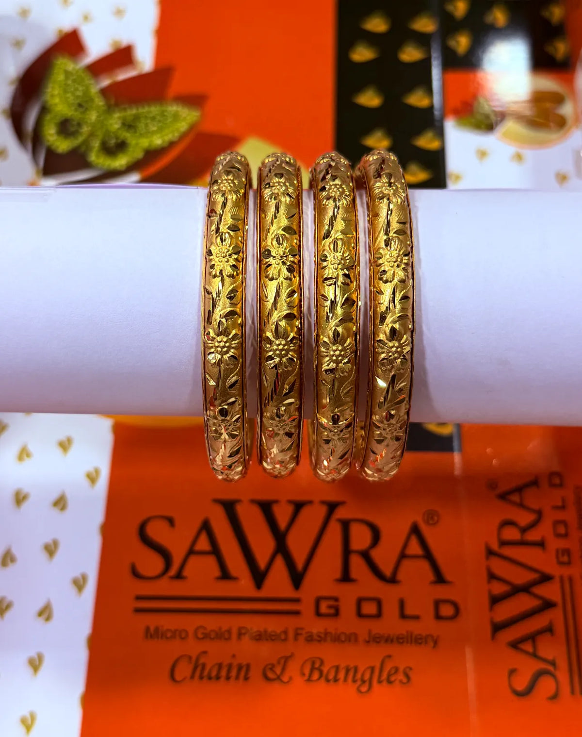 Four gold-plated bangles with intricate floral engravings, displayed on a white cylindrical stand in front of an orange SAWRA GOLD branded background.
