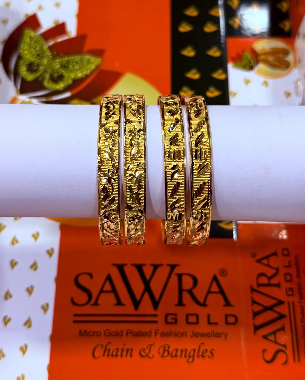 Four ornate gold bangles with intricate floral engravings displayed on a white cylindrical stand, with an orange SAWRA GOLD brand card in the foreground.