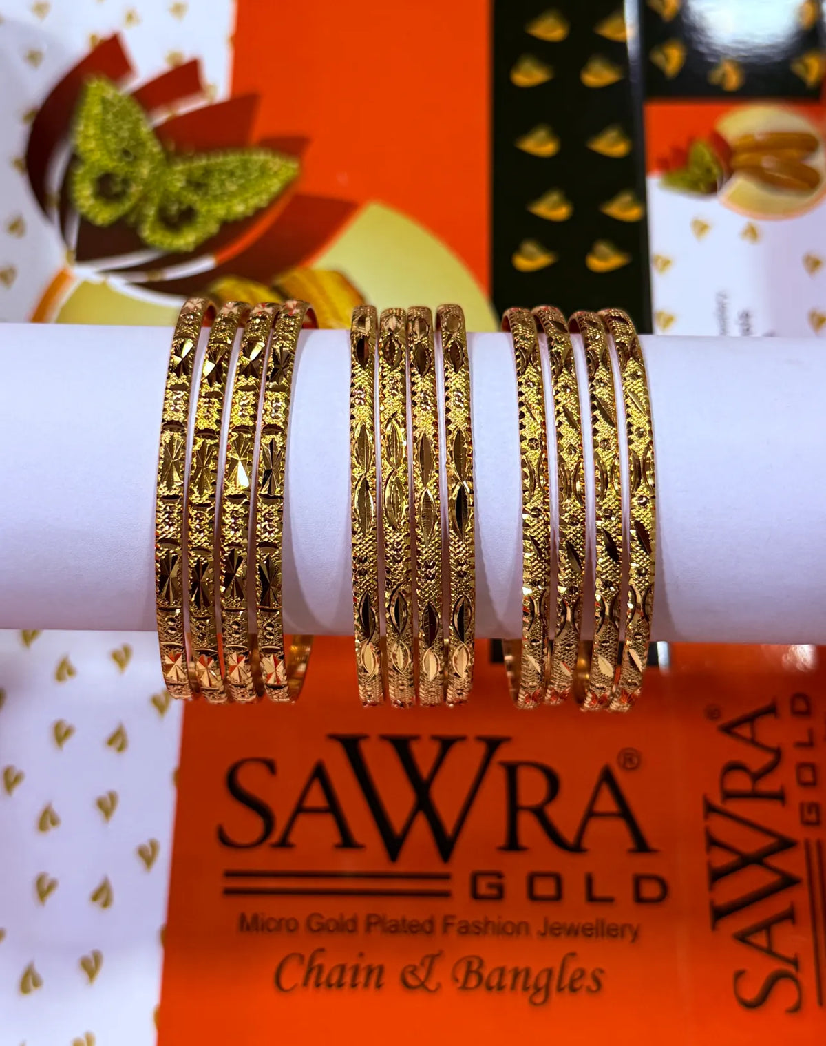 Three sets of gold-plated fashion bangles with intricate geometric and floral engravings, displayed on a white cylindrical stand. The background features branded orange and white packaging for SAWRA GOLD Chain & Bangles.