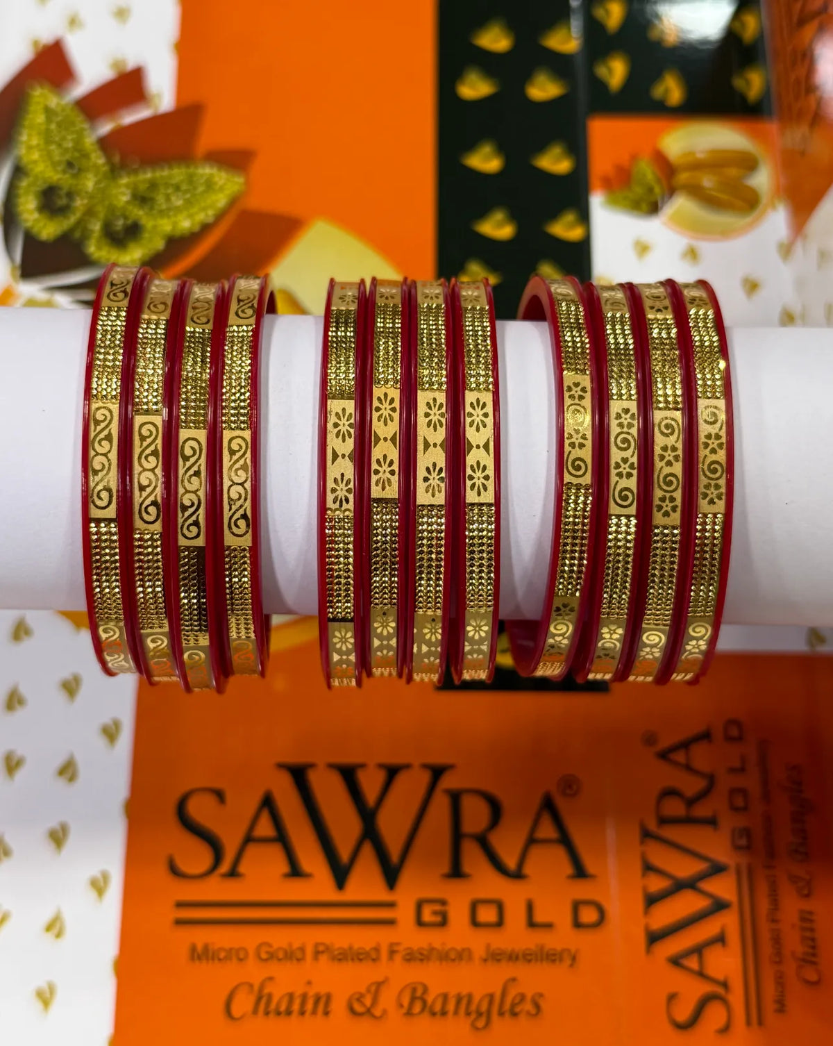 A set of red bangles with intricate gold-plated chain and floral patterns, displayed on a white cylindrical stand. The background features a branded orange and white box for SAWRA GOLD.